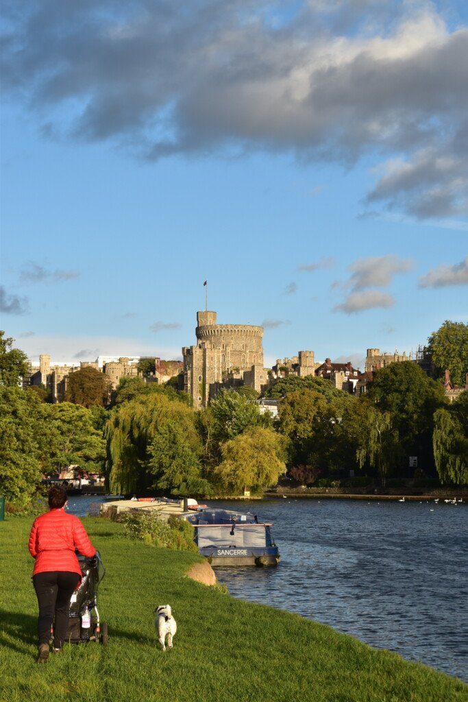 Hiker with pram marches through a field along the banks of the River Thames with Windsor Castle in the distance.