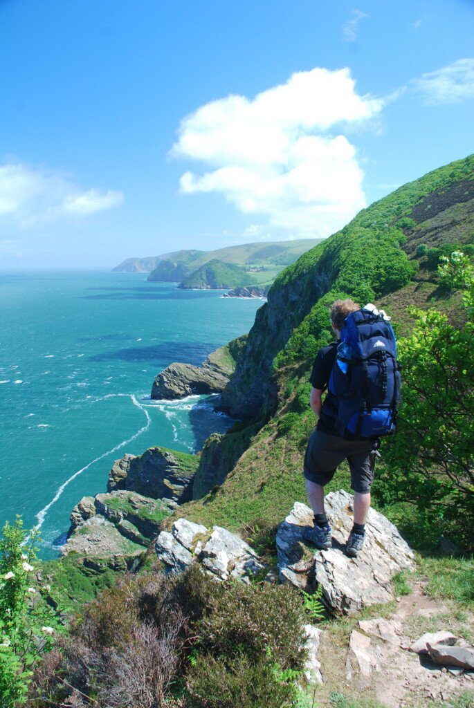 Lone hiker with big blue rucksack stares out over a turquoise sea from a rocky outcrop in North Devon
