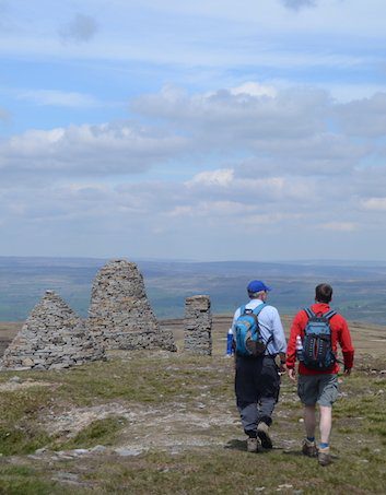 Two hikers walk away from camera past several large piles of stones known as the Nine Standards Rigg