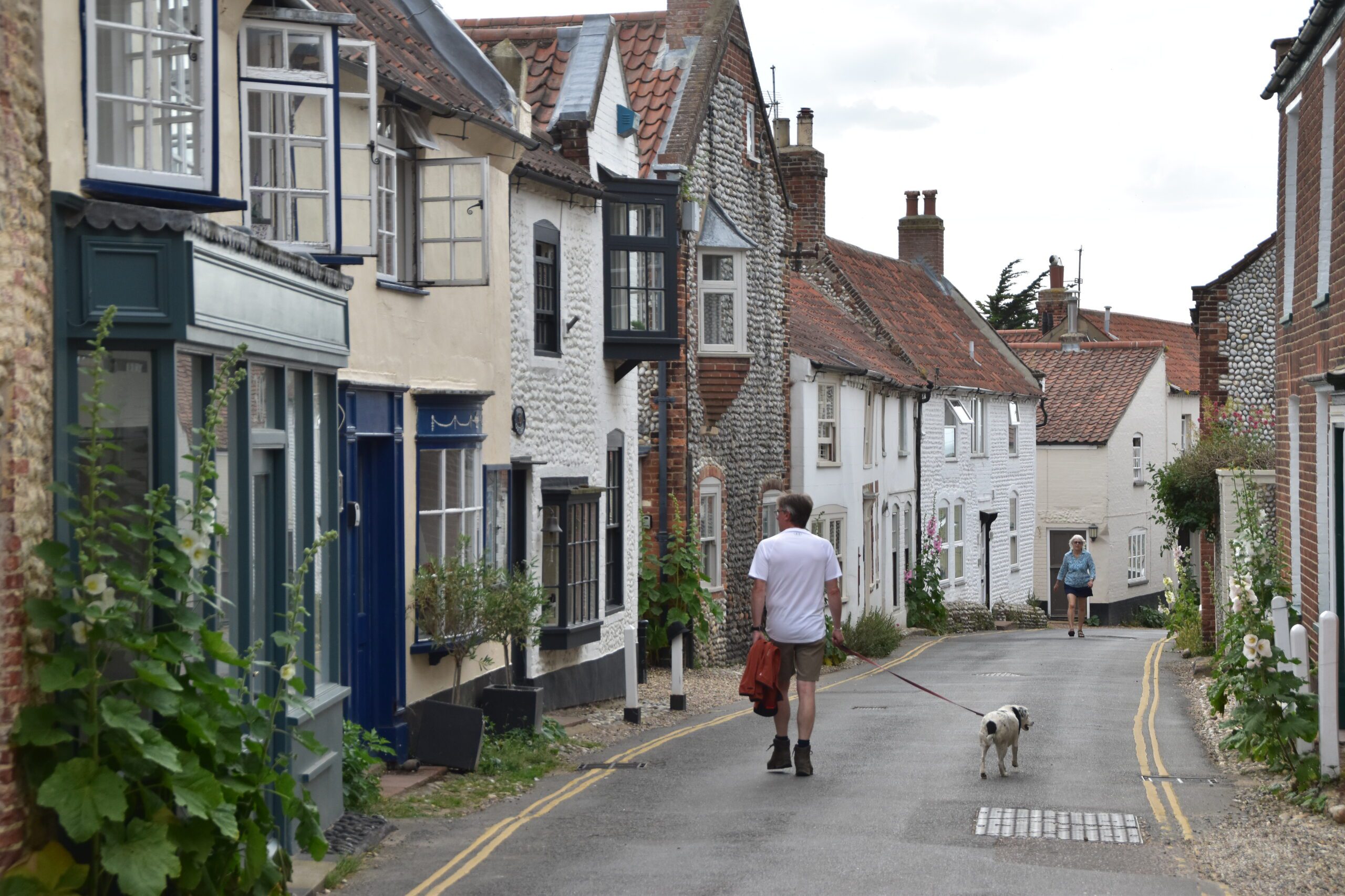 Hiker and Daisy walk down the main street in Blakeney, Norfolk, past lovely pebble cottages